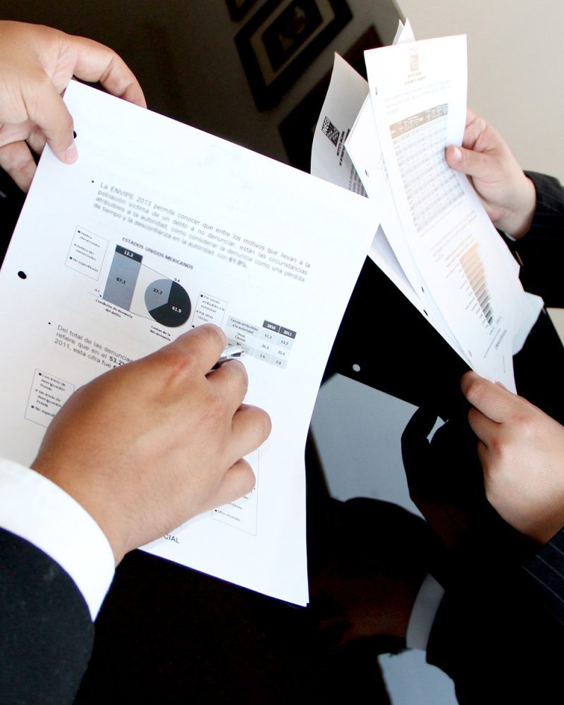 Close-up of two individuals in business attire holding and discussing printed documents with charts, graphs, and data in Spanish, indicating a professional meeting or financial review. One person is pointing at a pie chart with a pen.