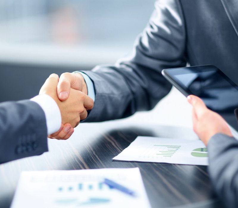 Two business professionals in suits shaking hands across a table, signifying a successful deal or agreement, while a third person holds a tablet and reviews charts and graphs on printed documents, representing a corporate or financial meeting.