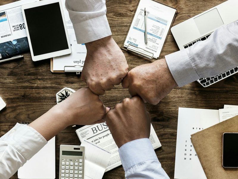 Four people performing a fist bump over a wooden office desk cluttered with business documents, a tablet, a laptop, a smartphone, a calculator, a coffee cup, and office supplies, symbolizing teamwork and collaboration in a professional setting.
